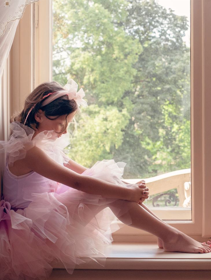 Dziewczynka w różowej opasce i sukience siedzi na parapecie okna, patrzy na swoje ręce i stopy