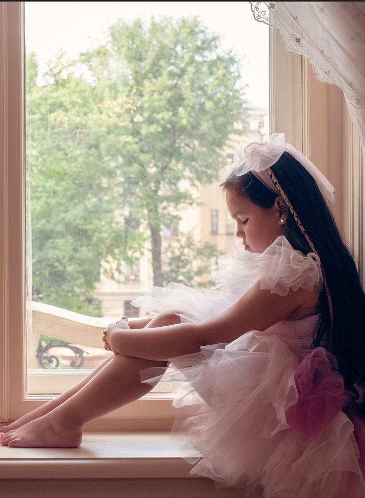 Dziewczynka w różowej opasce i sukience siedzi na parapecie okna i patrzy się na swoje ręce i nogi - sesja fotograficzna kampanii OKNO