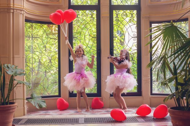 Dwie dziewczynki w różowych sukienkach tańczą i bawią się czerwonymi balonami w kształcie serc na tle okna - sesja kampanii OKNO