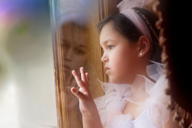 Kilkuletnia dziewczynka w różowej sukience i opasce, bohaterka kampanii OKNO patrzy przez okno