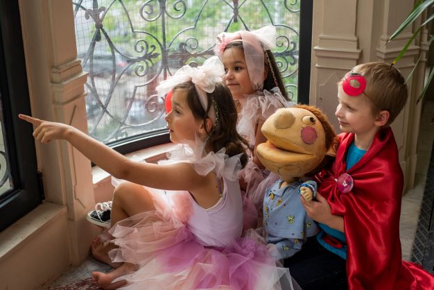 Dwie dziewczynki w różowych sukienkach i opaskach, chłopiec oraz Edek patrzą przez okno - sesja kampanii OKNO