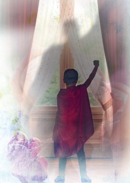 Chłopczyk w pelerynie superbohatera z balonami patrzy przez okno
