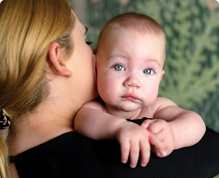 Opieka paliatywna mama Nadii trzyma Nadię na barku, która patrzy się bezpośrednio na kamerę raport 2024